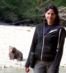 a woman standing next to a stream with a bear in the background